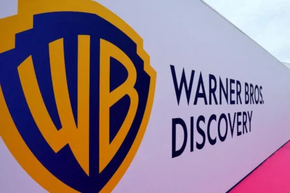 Logo di Warner Bros. Discovery su sfondo bianco con testo a destra, tappeto rosa e persone sullo sfondo