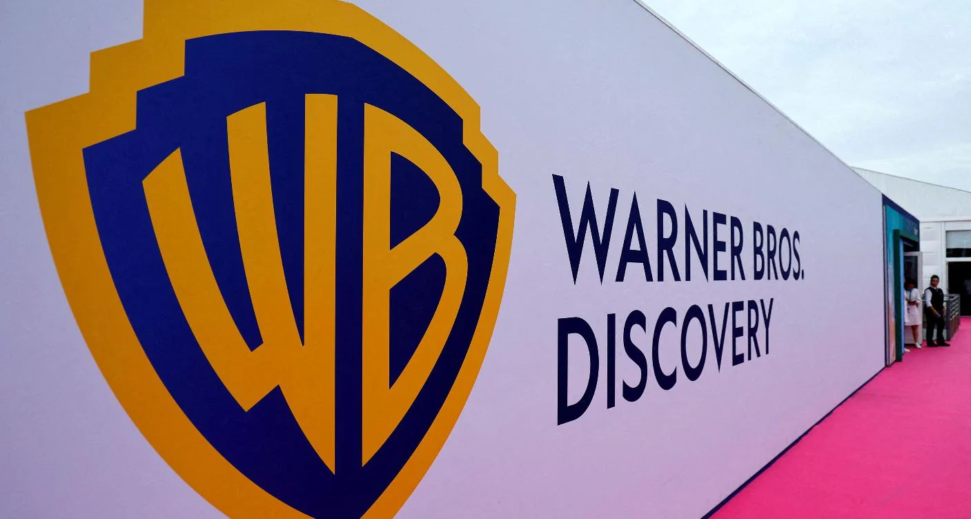 Logo di Warner Bros. Discovery su sfondo bianco con testo a destra, tappeto rosa e persone sullo sfondo